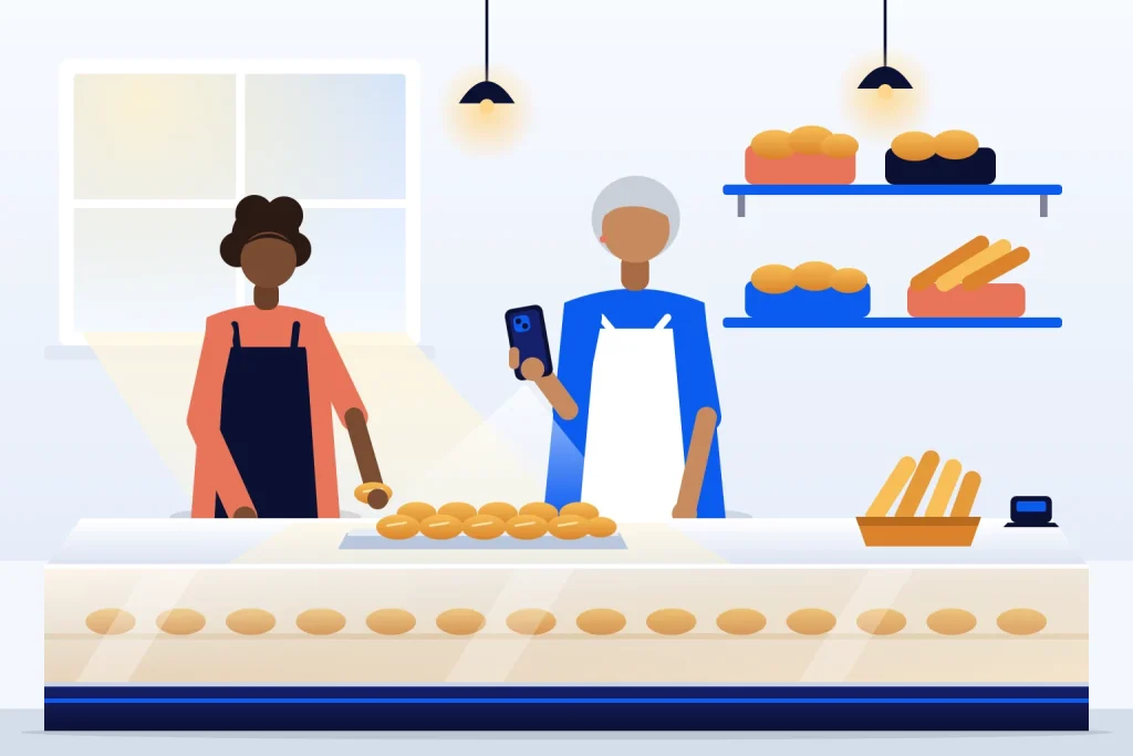 Dona de padaria filma bandeja de pães com o celular na vertical para criar anúncio em vídeo, enquanto funcionária arruma a vitrine
